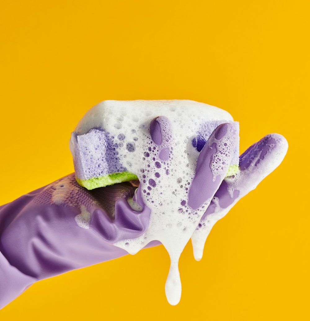 Purple cleaning glove holding a foamy sponge against a yellow background, from Shine Bright’s current website